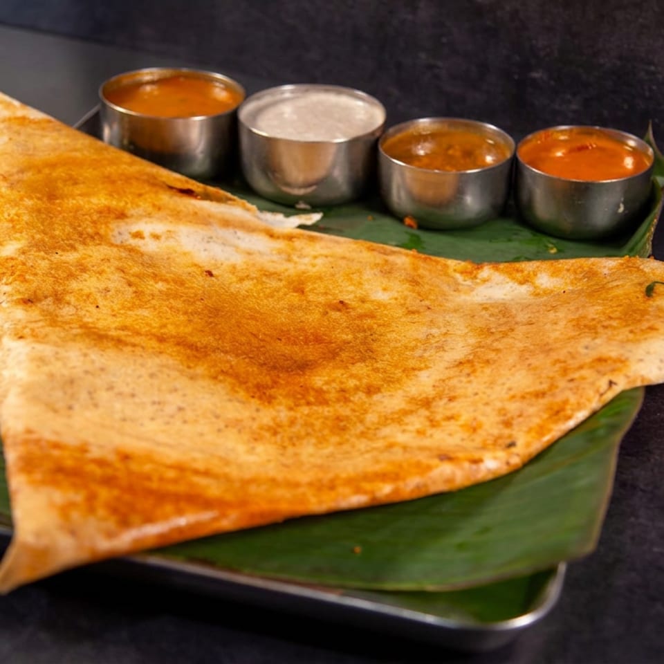 Crispy golden dosa served on a banana leaf with four metal bowls of sambar, chutney, and curry.
