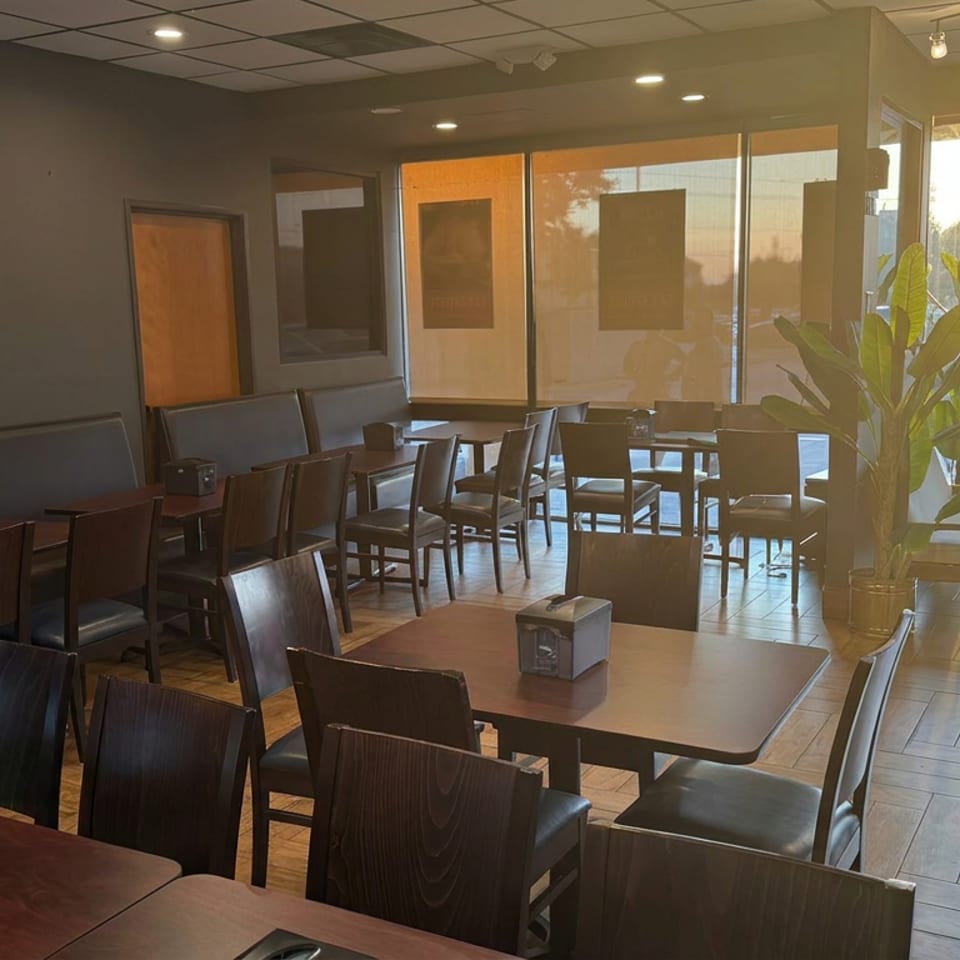Sunlit restaurant dining room with dark wooden tables, upholstered seating, large windows, and a tall potted plant.